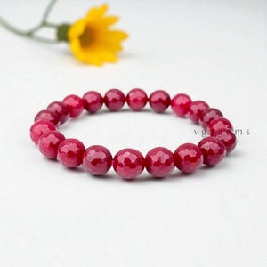 May include: A striking red gemstone bracelet. The bracelet features round, faceted beads, displaying a deep, rich hue. A yellow flower and green stem are in the background. The bracelet is a piece of jewellery.