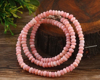 May include: A pink gemstone necklace with a faceted bead design. The necklace is made of small, round, light pink beads strung together. The clasp is silver. The necklace is displayed on a wooden surface.