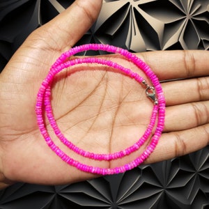 May include: A bright pink necklace made of small, circular beads. The necklace is shown on a hand, highlighting its colour and design. The clasp is silver. The background features a dark, geometric pattern.