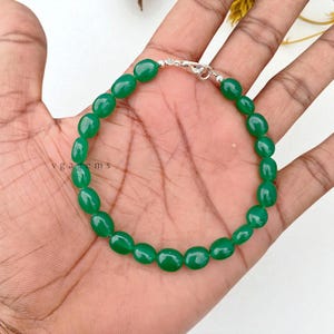 May include: A vibrant green gemstone bracelet featuring oval-shaped beads. The beads are strung together with a silver clasp. The bracelet is displayed on a hand, showcasing its color and design. The text "vgagems" is visible.