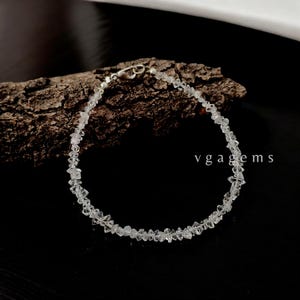 Pode incluir: Uma pulseira delicada com pequenas pedras preciosas claras de formato irregular, unidas. A pulseira tem um fecho prateado e é exibida sobre um fundo escuro com um pedaço de madeira. O texto "vgagems" é visível.