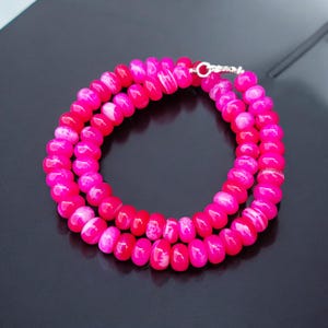 Puede incluir: Un collar de cuentas de ágata rosa y blanca con un cierre plateado. Las cuentas son redondas y tienen un acabado liso y pulido.