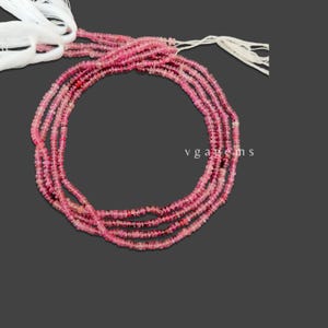May include: Strands of pink gemstone beads with white string tassels. The beads are small and round, with varying shades of pink. The background is a dark gray.