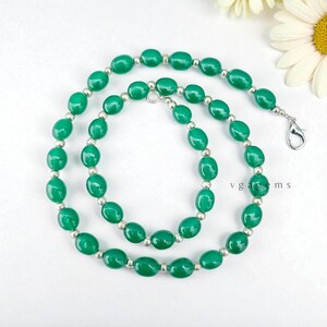 May include: A green beaded necklace with oval-shaped beads and silver-colored spacers. The necklace is displayed on a white surface with a flower in the background. The text "v g a g e m s" is visible.