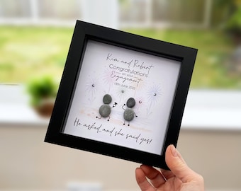 May include: A square black shadow box frame containing a white background with pebble art of two figures. The text reads: Kim and Robert, Congratulations on your Engagement, 18th June 2025, He asked... and she said yes! Fireworks illustrations are included.