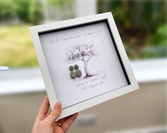 May include: A square white shadow box frame containing a pebble art piece. The artwork features two pebble figures on a swing under a printed tree, with the text On the Renewal of your Wedding Vows, Ashley & Robert Taylor, and 24th August 2026.