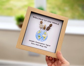 May include: A square light wood frame containing pebble art of a family of three standing on a globe illustration. The text reads Welcome to the World, Joseph Christopher Taylor, Born 14th May 2026. The figures are made of small brown and grey stones.