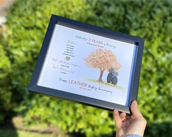 May include: A framed print celebrating a 3-year wedding anniversary. The artwork features a tree with a couple under it, with text that reads "Happy Leather Wedding Anniversary." The frame is black.