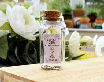 May include: A clear glass bottle with a cork stopper and a silver ribbon around the neck. Inside is a rectangular tag with the text On Our 25th Anniversary, Lisa & Alex, 28.09.01, and a small crystal accent. The bottle contains metallic confetti.