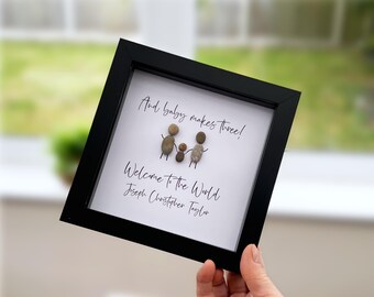 May include: A square black frame containing white paper with pebble art depicting three figures. The text reads: And baby makes three! Welcome to the World Joseph Christopher Taylor. The figures are made of small, smooth, grey and brown stones.