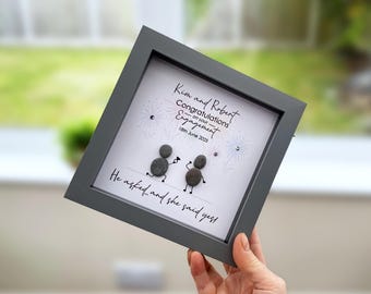 May include: A square dark gray shadow box frame containing pebble art of two figures. The text reads: Kim and Robert, Congratulations on your Engagement, 18th June 2025, He asked... and she said yes. The background features illustrated fireworks.