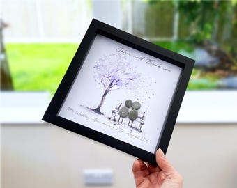 May include: A square black frame containing a white print of a tree and a bench with four small gray pebbles arranged to represent figures. The text reads John and Barbara and 70th Wedding Anniversary 24th August 1956 in black script.