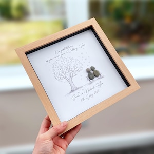 May include: A square light wood frame containing a white print of a tree with heart-shaped leaves and two figures sitting on a bench made of grey pebbles. The text reads: Congratulations as you renew your Wedding Vows, Sarah & Michael Taylor, 11th July 2026.