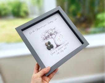 May include: A square gray framed wall art piece featuring pebble art of two figures sitting under a tree. The text reads On the Renewal of your Wedding Vows, Sarah & Michael Roberts, 17th May 2026. The tree has small gold heart accents.