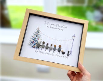 May include: A framed pebble art piece featuring a family of seven figures and two pets standing under a string of lights next to a Christmas tree and a street lamp. The text reads 'Tis the season to be jolly!' and 'The Robinson Family' with names below.