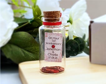 May include: A clear glass bottle with a cork stopper and a red ribbon around the neck. A rectangular label features the text "On Your 1st Anniversary Jonathan & Charlie 27.08.25" with a red heart accent. Red heart-shaped confetti fills the bottom.