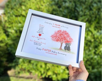 May include: A framed print celebrating a 1-year wedding anniversary. The artwork features a red tree, two pebble figures, and text that reads "Happy Paper Wedding Anniversary" and the date. The frame is white.