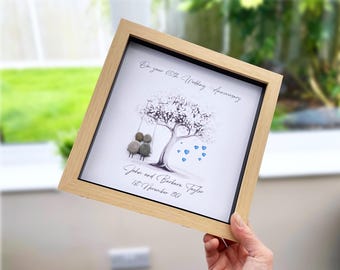 May include: A square light wood frame containing a pebble art piece. It features a tree with a swing, two pebble figures, and blue heart accents. Text reads: On your 65th Wedding Anniversary, John and Barbara Taylor, 1st November 1961.