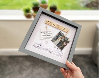 May include: A gray framed wedding keepsake with the word "MARRIED" spelled out in wooden letter tiles. The frame holds a photo, a congratulatory note, and decorative elements. A hand holds the frame.