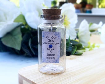 May include: A clear glass bottle with a cork stopper containing a small rectangular tag that reads On Our 2nd Anniversary Mark & Sarah 18.06.24 with a blue rhinestone accent. The bottle is filled with small white circular confetti pieces.