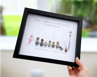 May include: A black framed wall art piece featuring pebble art figures representing a family of seven, a dog, and a cat. The print includes a snowman, a lamp post, and the text: Family - Together is our favourite place to be, The Robinson Family.