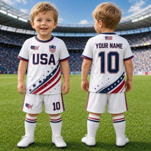 A young boy wearing a white soccer uniform with red, white, and blue colors, standing on a soccer field.