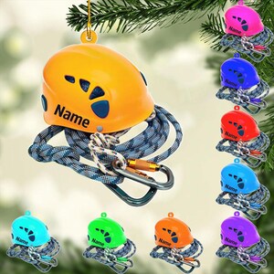 May include: A yellow climbing helmet ornament with the word "Name" printed on it, paired with a blue and gray rope, a carabiner, and a gold-colored hanging loop. Additional ornaments in various colors are also shown.