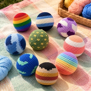 May include: A collection of colorful, handmade crochet balls. The balls feature various designs, including rainbow stripes, geometric patterns, and a globe. The balls are arranged on a striped blanket, with yarn in a basket in the background.