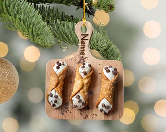 May include: A wooden Christmas ornament shaped like a cutting board, featuring three cannoli pastries with chocolate chips. The ornament is hanging from a gold string, with the word "Name" engraved on the handle.