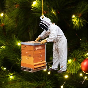 May include: A Christmas ornament featuring a beekeeper inspecting a wooden beehive. The hive is orange and brown with the name "Julian" on top. The beekeeper wears a white suit, gloves, and a protective hood. Bees are depicted on the hive.
