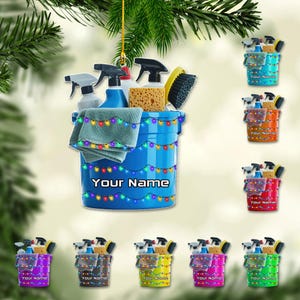 May include: A blue bucket-shaped ornament filled with cleaning supplies, including spray bottles, a sponge, and a brush. The ornament is decorated with colorful string lights and the text "Your Name". Additional ornaments in various colors are also shown.