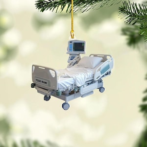 May include: A miniature hospital bed ornament with a monitor and the text "YOUR NAME". The bed has white linens and is suspended by a gold string. The ornament is a medical-themed decoration.