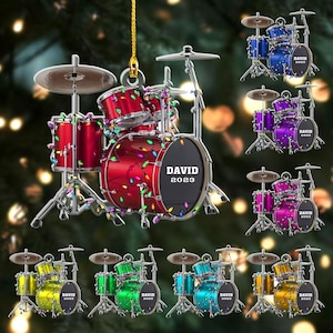 May include: A red drum set ornament with the text "DAVID 2023" on the bass drum. The ornament is decorated with colorful string lights. Additional drum set ornaments in blue, purple, pink, yellow, green, and teal are also shown.