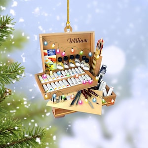 May include: Wooden artist's box ornament with the name "William" engraved on the lid. The box is filled with paint tubes, brushes, paint bottles, and other art supplies. A string of colorful lights decorates the box, suggesting a festive theme.