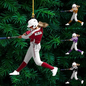 May include: A softball player ornament in a batting pose, wearing a white helmet and uniform with the name "ALISA" and the number "18". The ornament is red, white, and gray. Additional ornaments in orange, purple, and black are also shown.