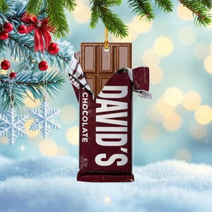 May include: A chocolate bar ornament with the text "DAVID'S CHOCOLATE" in white on a maroon wrapper. The chocolate bar is brown and partially unwrapped, revealing the chocolate squares. The ornament is hanging from a gold string.