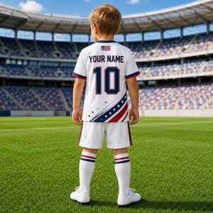A young boy wearing a white soccer jersey with the text "YOUR NAME 10" and an American flag design on the back, standing on a soccer field.