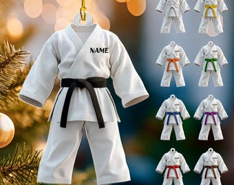 May include: A white karate uniform ornament with a black belt and the word "NAME" printed on the chest. Additional ornaments feature white uniforms with colored belts: yellow, orange, green, blue, purple, red, and brown. The ornament hangs from a gold string.