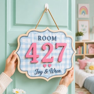 May include: A decorative wall sign with a light blue and white gingham pattern, featuring the text ROOM 427 in large pink numbers, two small pink hearts, and the names Ivy & Wren in dark blue script, suspended by a tan rope from a white hook.