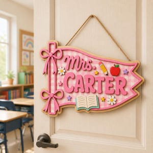 May include: A wooden pennant-shaped door sign featuring a pink and white gingham background. The text reads Mrs. CARTER in pink letters. Decorative elements include a pencil, a red apple, an open book, daisies, and stars, with two pink bows.
