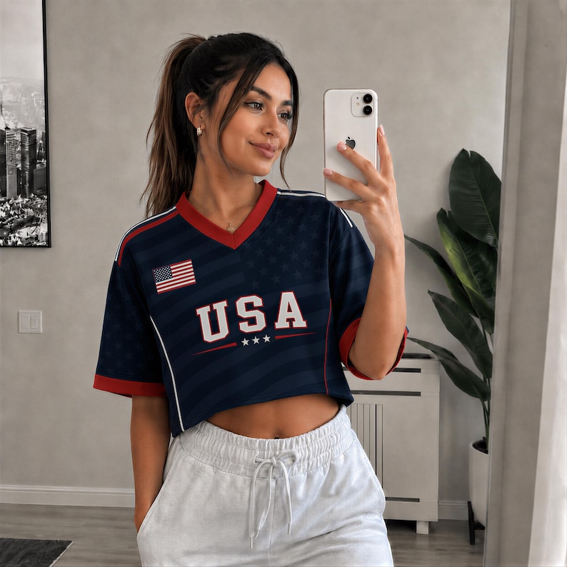 A woman wearing a blue USA jersey and white sweatpants is taking a selfie with a smartphone in a room with a plant and a framed picture on the wall.