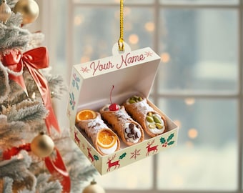 May include: A decorative ornament shaped like a box of cannoli pastries. The box is cream-colored with red reindeer and green holly designs. The cannoli are filled with various toppings, including orange slices, chocolate chips, and pistachios. The text "Your Name" is printed on the box.