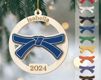 May include: Wooden Christmas ornament with a blue belt design, the name "Isabella" and the year "2024" engraved. A selection of colored belt designs are shown on the right. The ornament hangs from a gold string.