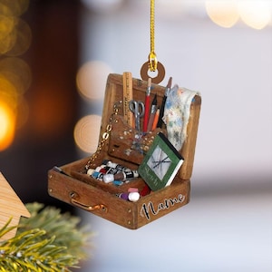 May include: A miniature wooden artist's toolbox ornament. The open box reveals tiny paint tubes, brushes, scissors, and a sketchbook. The ornament is suspended by a gold cord and features the handwritten word "Name" on the front.