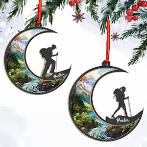 May include: Two wooden crescent moon ornaments with a hiker silhouette and a landscape scene. The ornaments have a red ribbon for hanging and the name "Parker" is printed on the bottom. The landscape features a river, mountains, and trees.