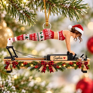 May include: A Christmas ornament featuring a figure in a Santa hat performing a plank on a Pilates reformer machine. The figure wears red and white patterned leggings and a matching top. The reformer base is decorated with pine branches, red berries, and plaid bows, with the name Sophia and a red heart printed on the side.