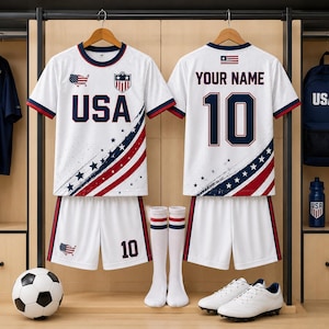A pair of white soccer uniforms with red and blue stripes, displaying the text "USA" and the number "10" on the back. The uniforms are hanging on a rack, along with a pair of white socks and a soccer ball.