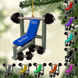 May include: A blue weight bench ornament with the name "Matthew" and the year "2023" printed on it. The ornament has black weights and a green frame. Additional ornaments with different colors are also shown.