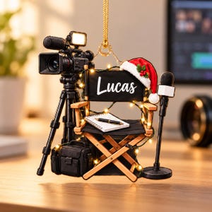 May include: A hanging ornament featuring a black director's chair with the name Lucas printed in white, a Santa hat, a video camera on a tripod, a microphone, and a notebook with a pen. The set is decorated with small warm white string lights.