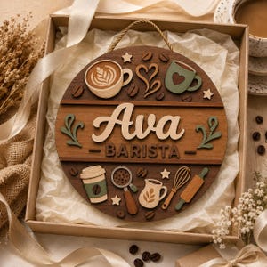 May include: A circular wooden wall sign featuring the name Ava and the word BARISTA in raised lettering. The design includes layered cutouts of coffee cups, a portafilter, a whisk, a rolling pin, and coffee beans in shades of brown and green.